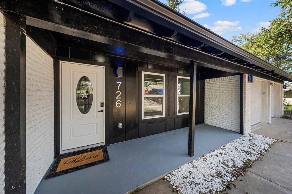 Covered porch entrance featuring painted brick masonry, dark exterior trim, a single-panel door with an oval window insert, and integrated exterior lighting