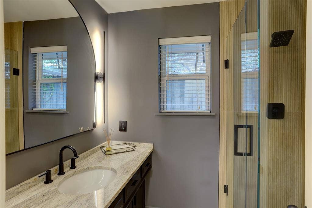 Vanity featuring a stone countertop with an integrated undermount sink, matte black widespread faucet, arched backlit mirror, and glass-enclosed shower with textured wall tile and matte black fixtures
