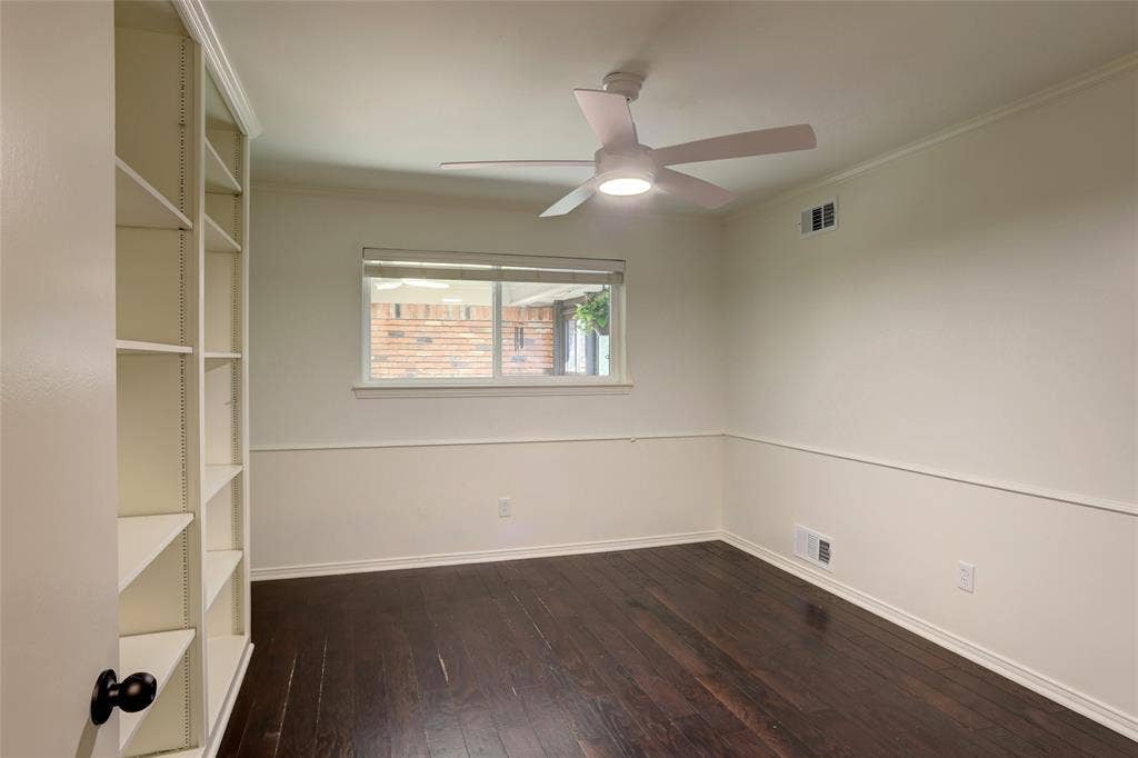Interior room featuring dark wood-finish flooring, built-in shelving, white wainscoting, and a ceiling fan with integrated lighting