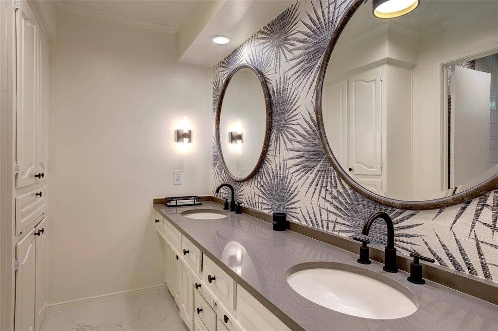 Dual-sink vanity featuring gray solid-surface countertops, white cabinetry with integrated storage, matte black widespread faucets, round mirrors, and patterned wall covering