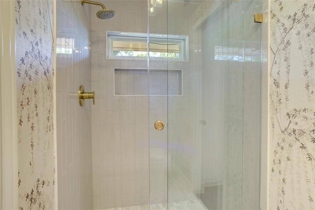Walk-in shower featuring a frameless glass door, light-toned wall tile, a built-in recessed storage niche, a horizontal transom window, and gold-finish hardware