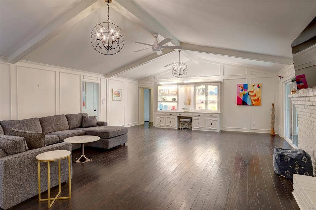 Vaulted ceiling with exposed beams, wood-finish flooring, built-in cabinetry with glass-front shelving, and a painted brick fireplace surround