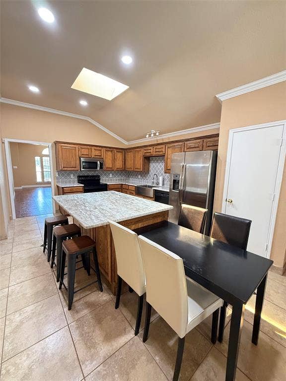 Kitchen featuring wood cabinetry, stone-finish countertops, a central island with integrated breakfast bar, a geometric-pattern tile backsplash, and a vaulted ceiling with a skylight