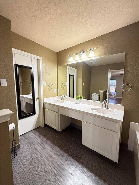 Dual-sink vanity with white cabinetry, light-toned countertop, large wall-mounted mirror, four-light vanity fixture, and wood-finish flooring