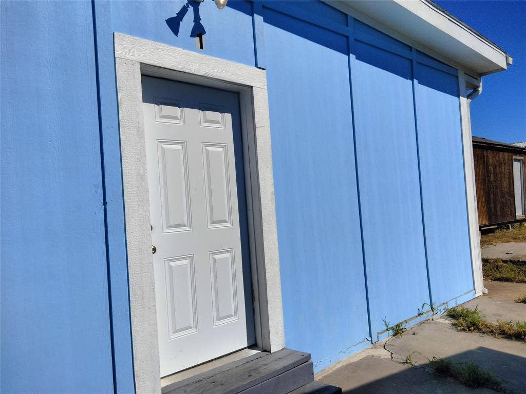 Exterior elevation featuring blue board-and-batten siding, a white four-panel entry door with thick casing, and a concrete patio