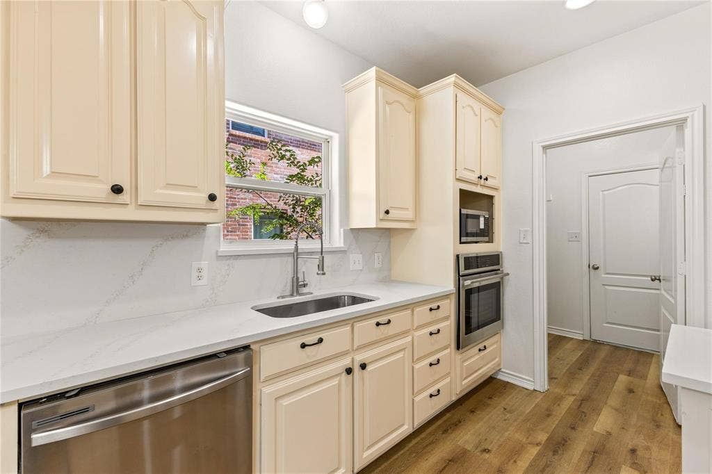 Kitchen featuring light-toned cabinetry, solid surface countertops with matching backsplash, stainless steel sink with high-arch faucet, integrated wall oven and microwave, and wood-finish flooring