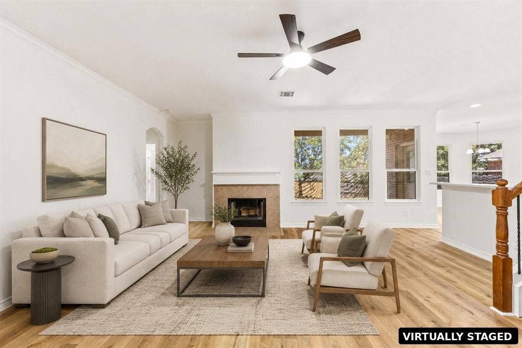 Living area featuring wood-finish flooring, a fireplace with a neutral tile surround, a ceiling fan with integrated lighting, and multiple tall windows