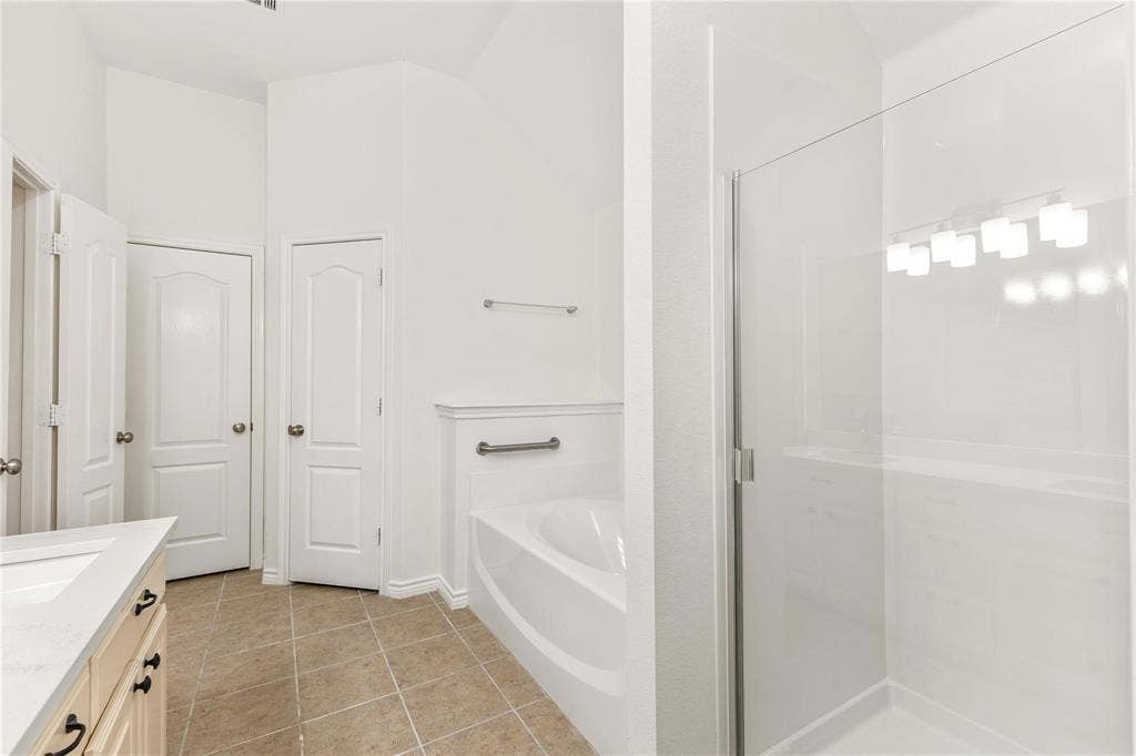 Bathroom featuring a built-in soaking tub, separate glass-enclosed shower, neutral-tone tile flooring, and light-toned cabinetry with a solid-surface countertop