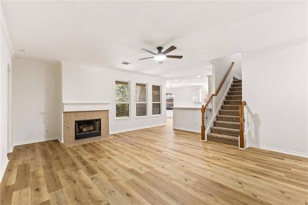 Open-concept area featuring wood-finish flooring, a fireplace with a stone-finish surround, triple-pane windows, a ceiling fan, and a staircase with a wood handrail