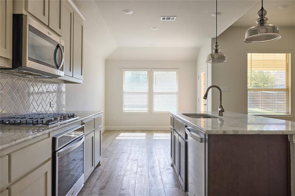 Kitchen featuring stone countertops, a center island with undermount sink, herringbone-patterned tile backsplash, stainless steel appliances, and wood-finish flooring