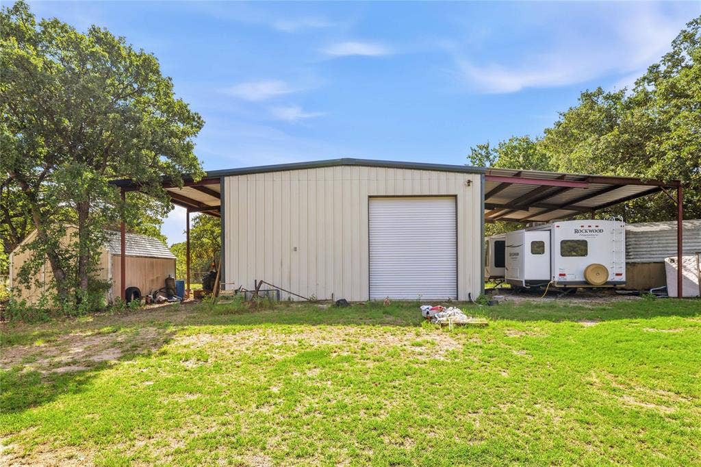 5540 County Road 334, Blanket, TX 76432 Metal-sided workshop featuring a roll-up garage door, expansive side-covered storage, and a detached secondary storage shed situated on a grassy lot with mature trees