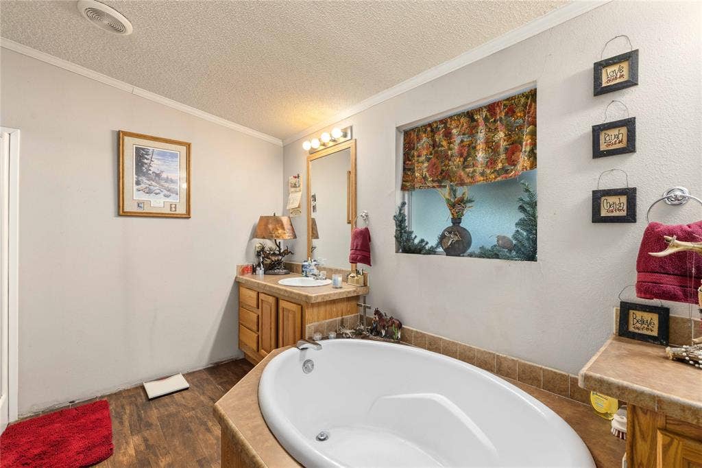 5540 County Road 334, Blanket, TX 76432 Oval soaking tub with tiled surround, wood-finish cabinetry, vanity mirror with overhead light fixture, and wood-finish flooring