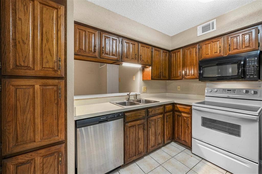 14800 Enterprise Drive, #22D, Farmers Branch, TX 75234 Kitchen featuring stained wood cabinetry, solid-surface countertops, stainless steel dishwasher, built-in microwave, and tile flooring