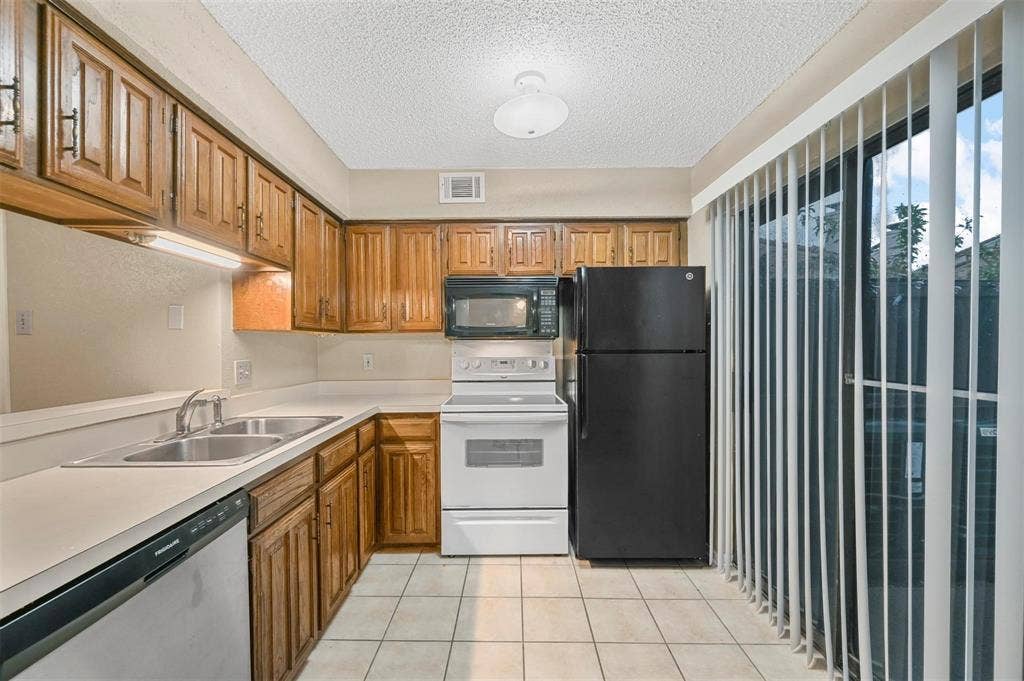 14800 Enterprise Drive, #22D, Farmers Branch, TX 75234 Kitchen featuring wood cabinetry, ceramic tile flooring, solid-surface countertops, dual-basin stainless steel sink, and sliding glass patio door