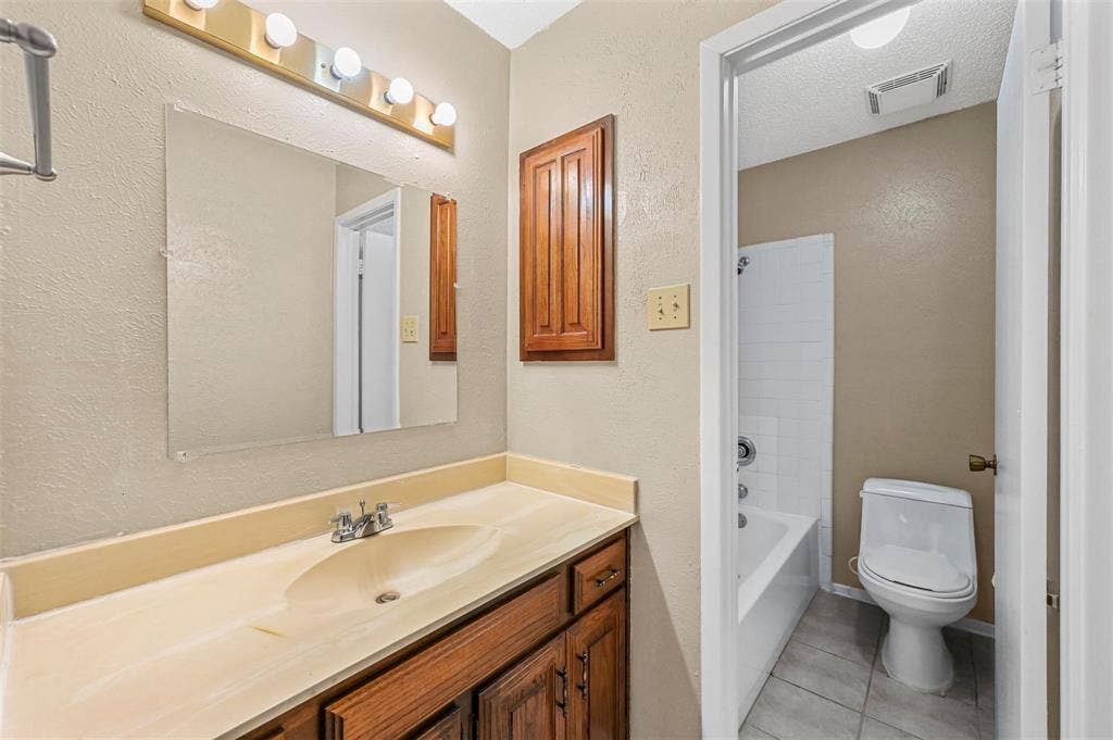 14800 Enterprise Drive, #22D, Farmers Branch, TX 75234 Bathroom featuring a vanity with an integrated basin, wood-tone cabinetry, wall-mounted medicine cabinet, four-bulb vanity light, and a tub-shower combination with tile surround