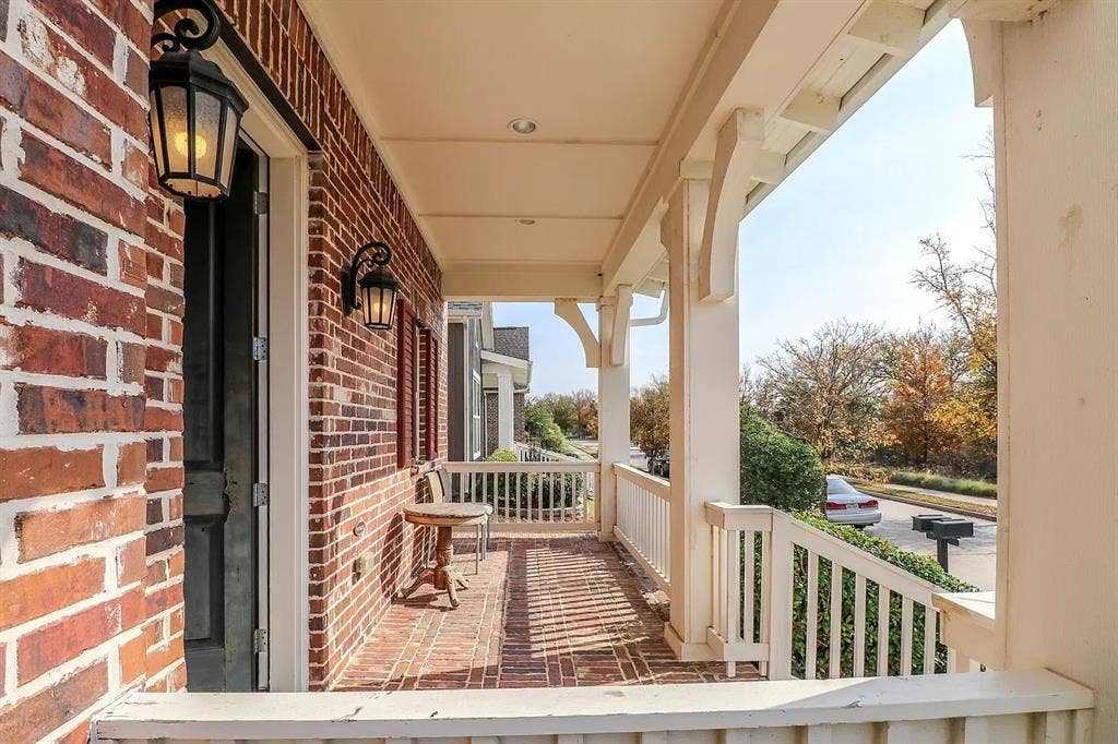Covered front porch featuring red brick flooring, decorative corbel brackets, and wall-mounted lantern-style exterior lighting