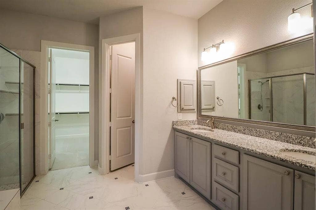 Bathroom featuring a dual-sink vanity with stone-patterned countertop, grey shaker-style cabinetry, framed vanity mirror, glass-enclosed shower, and large-format tile flooring
