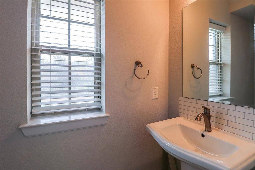 Pedestal sink with a metal faucet, subway tile backsplash, wall-mounted towel ring, and window with slat blinds