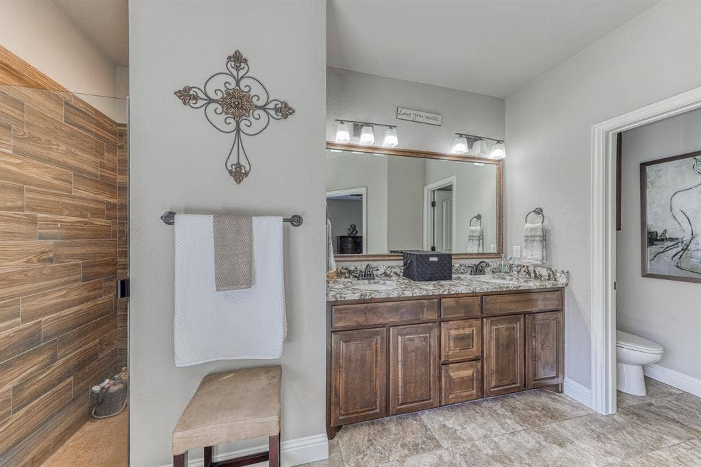 128 Breeders Drive, Willow Park, TX 76087 Double vanity featuring stained wood cabinetry, stone-patterned countertops, dual undermount sinks, and full-width wall mirror, complemented by a walk-in shower with wood-finish tile wall surfacing