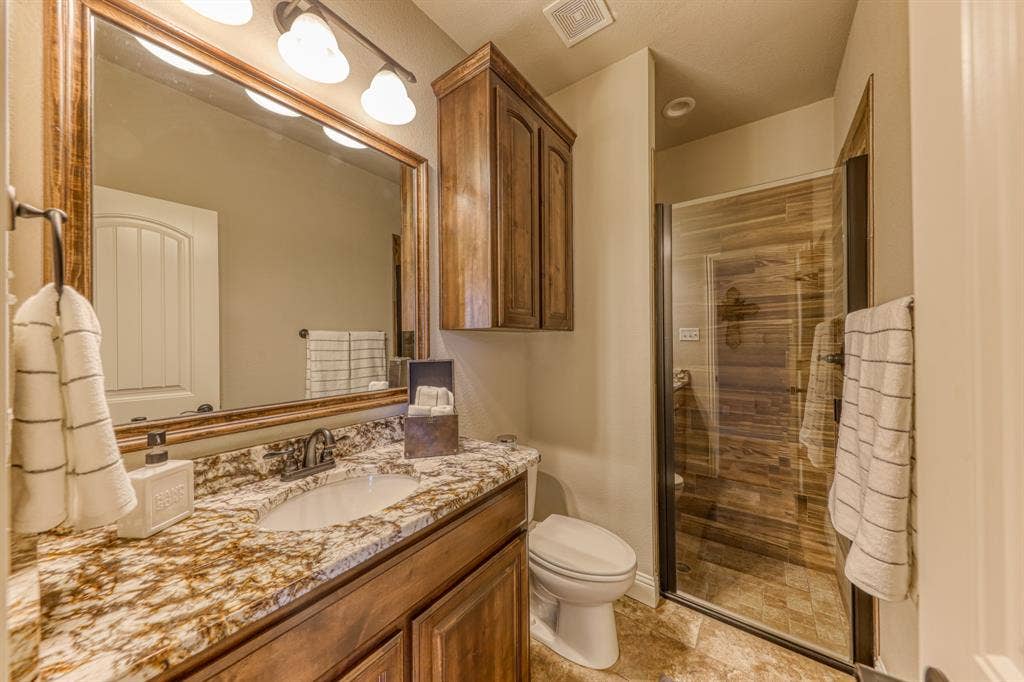 128 Breeders Drive, Willow Park, TX 76087 Bathroom featuring stone-finish countertop, dark wood-finish cabinetry, framed vanity mirror, and glass-enclosed shower with tile surround