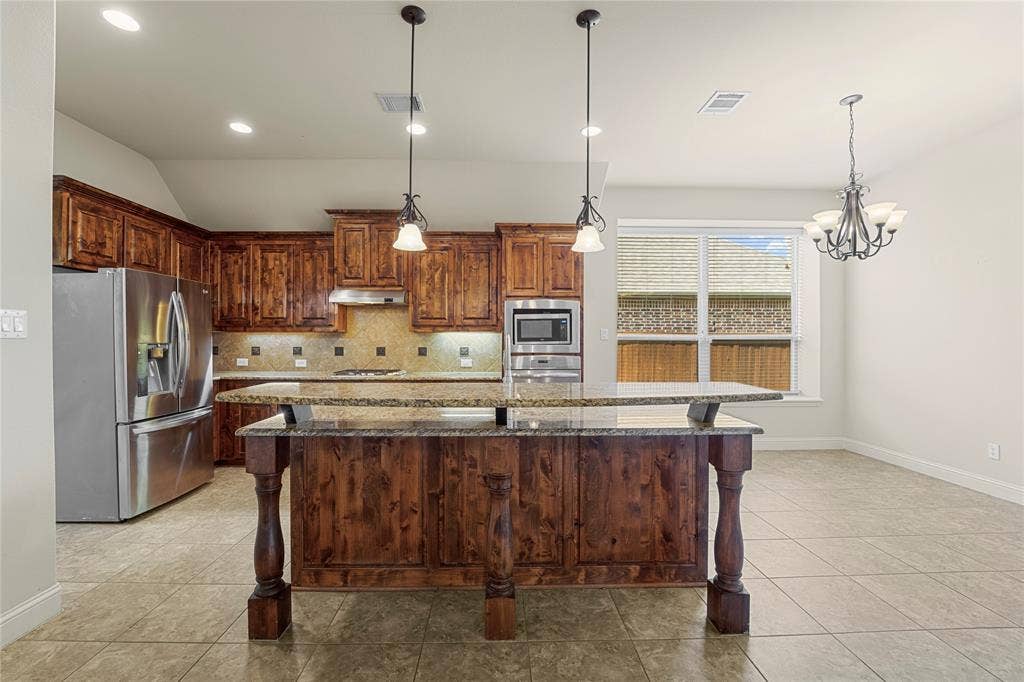 Kitchen featuring a center island with a stone-finish countertop, stained wood cabinetry, stainless steel appliances, dual pendant lighting, and tile flooring