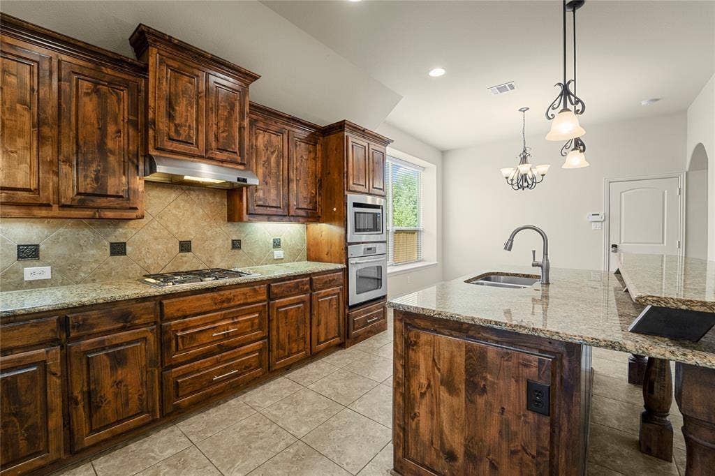 Kitchen featuring stained wood cabinetry, stone-surface countertops, tile backsplash, built-in double wall ovens, and a center island with a stainless steel sink