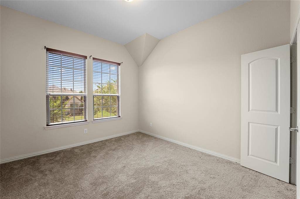 Vaulted ceiling room featuring neutral carpeting, two dual-pane windows with horizontal blinds, and a solid-core interior door