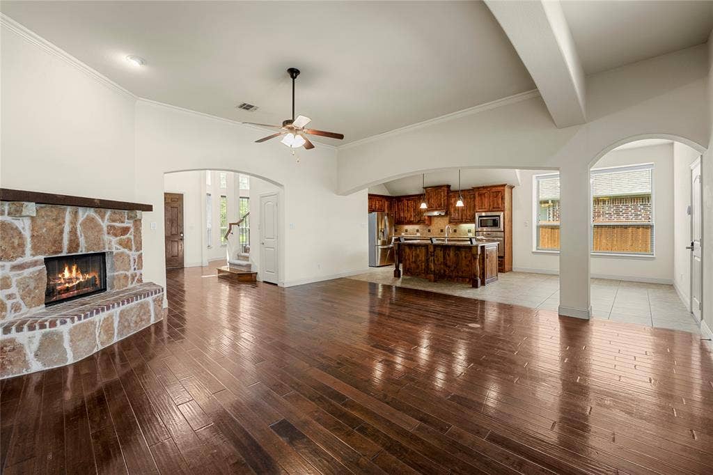 Open-concept interior featuring wood-finish flooring, a stone-faced fireplace with a timber mantel, arched architectural detailing, a central ceiling fan, and a kitchen equipped with wooden cabinetry and an island