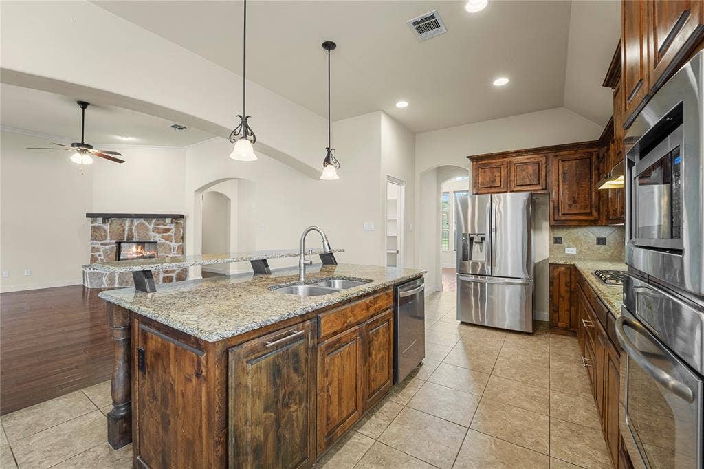 Kitchen featuring stone-finish countertops, wood cabinetry, stainless steel appliances, dual-basin sink, and tile flooring, with an additional stone-finish fireplace visible