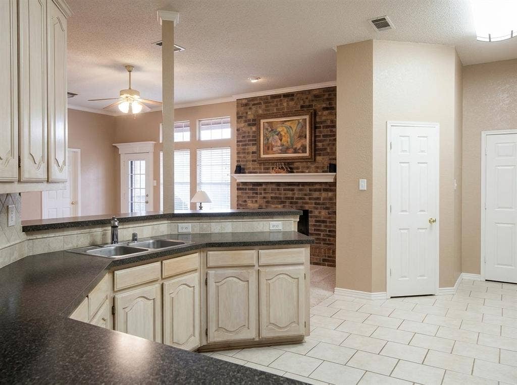 2306 Summit Drive, McKinney, TX 75071 Kitchen featuring off-white cabinetry, dark countertops, a double-basin stainless steel sink, and tile flooring, with a brick fireplace surround