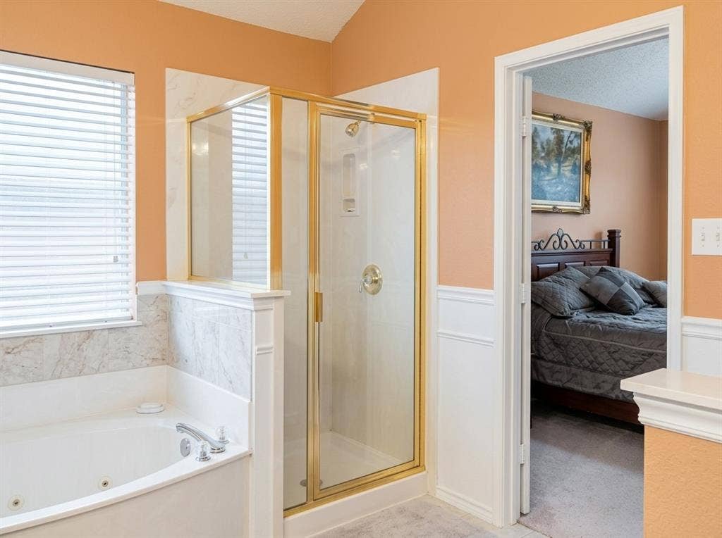 2306 Summit Drive, McKinney, TX 75071 Jetted soaking tub with tile surround, glass-enclosed shower with gold-tone frame, and white wainscoting