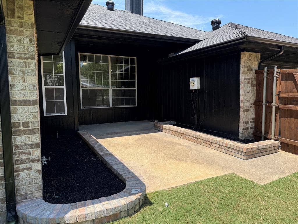 2400 Waterloo Lane, Mesquite, TX 75181 Rear exterior featuring a concrete patio, brick-trimmed landscaping beds, dark-finished vertical siding, a multi-pane grid window, and a wooden privacy fence
