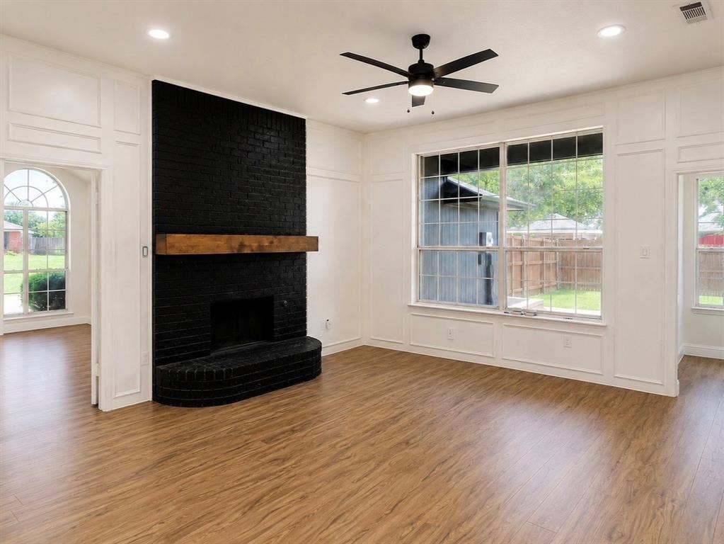 2400 Waterloo Lane, Mesquite, TX 75181 Black painted brick fireplace with a natural wood mantel, wood-finish flooring, grid-pattern windows, wainscoting wall paneling, and a ceiling-mounted light fixture with fan