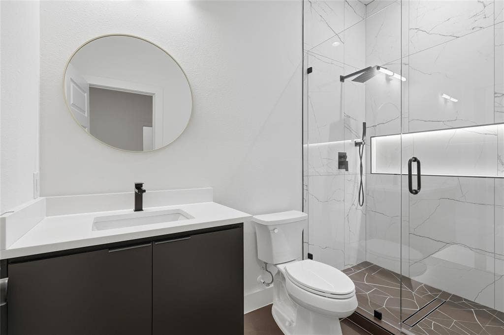 1834 Gallagher Street, Dallas, TX 75212 Modern bathroom featuring a dark cabinetry vanity with a white countertop, circular wall-mounted mirror, walk-in shower with large-format tile surround, integrated recessed lighting, and matte black fixtures