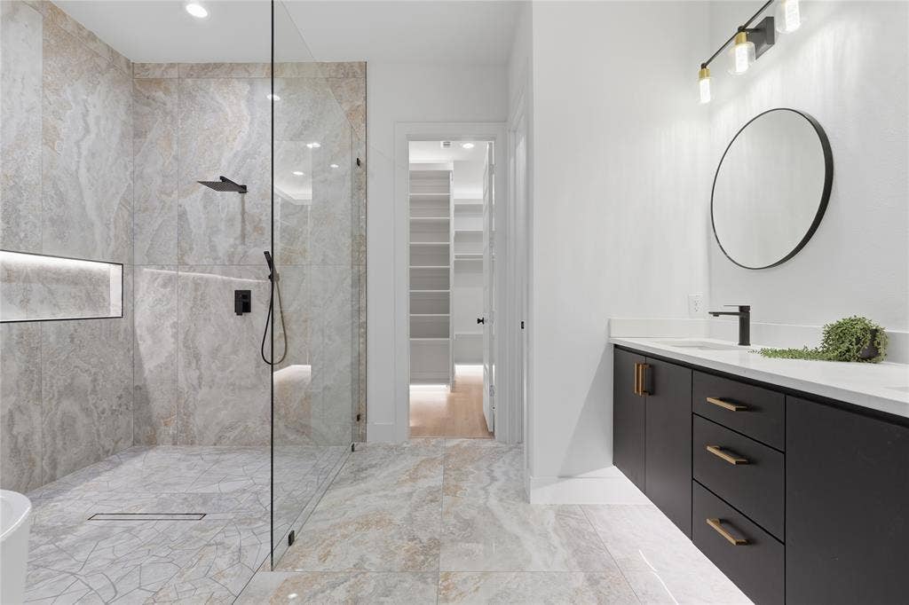 1834 Gallagher Street, Dallas, TX 75212 Walk-in shower with floor-to-ceiling tile, integrated recessed shelving, frameless glass enclosure, matte black rainfall shower fixture, and vanity with dark cabinetry, stone countertop, and circular mirror