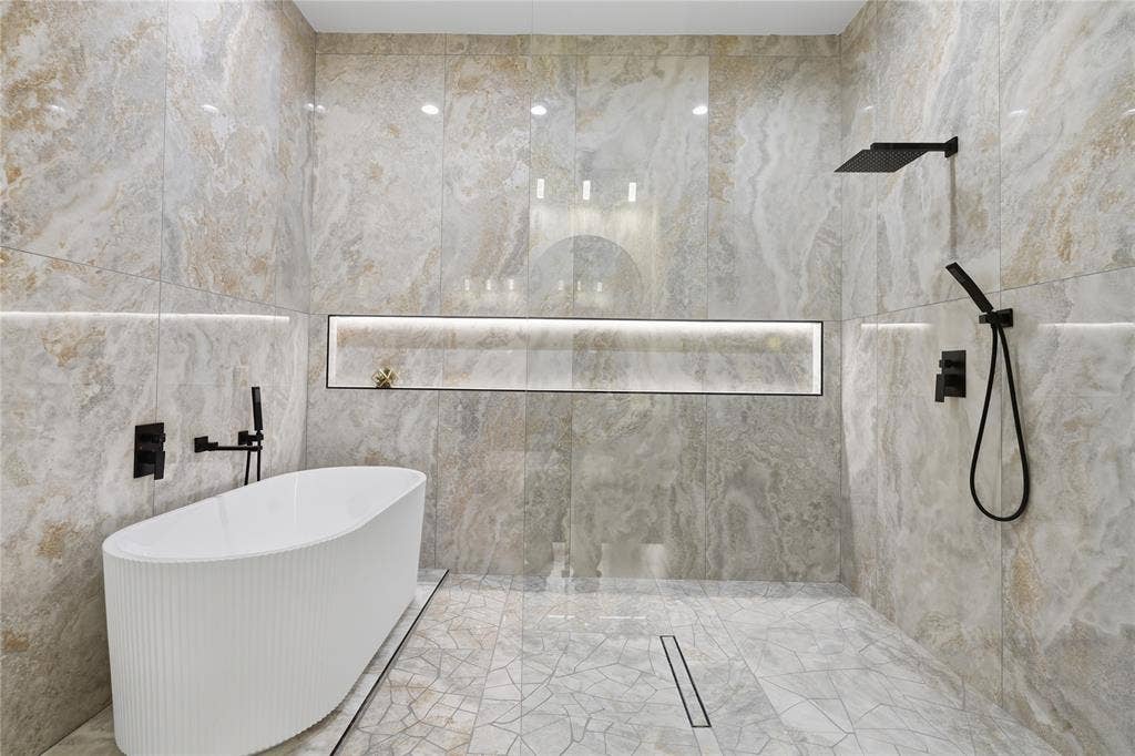 1834 Gallagher Street, Dallas, TX 75212 Freestanding white soaking tub, floor-to-ceiling large-format stone tile walls, matte black rainfall shower head with handheld attachment, integrated wall-recessed shelf with lighting, and linear floor drain
