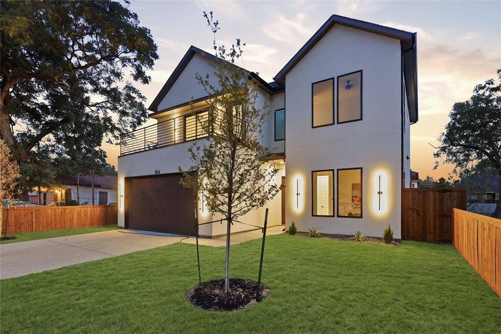 1834 Gallagher Street, Dallas, TX 75212 Two-story residence featuring a smooth stucco exterior, a second-level balcony with horizontal metal railing, a concrete driveway, modern wall-mounted exterior lighting, and a wood privacy fence