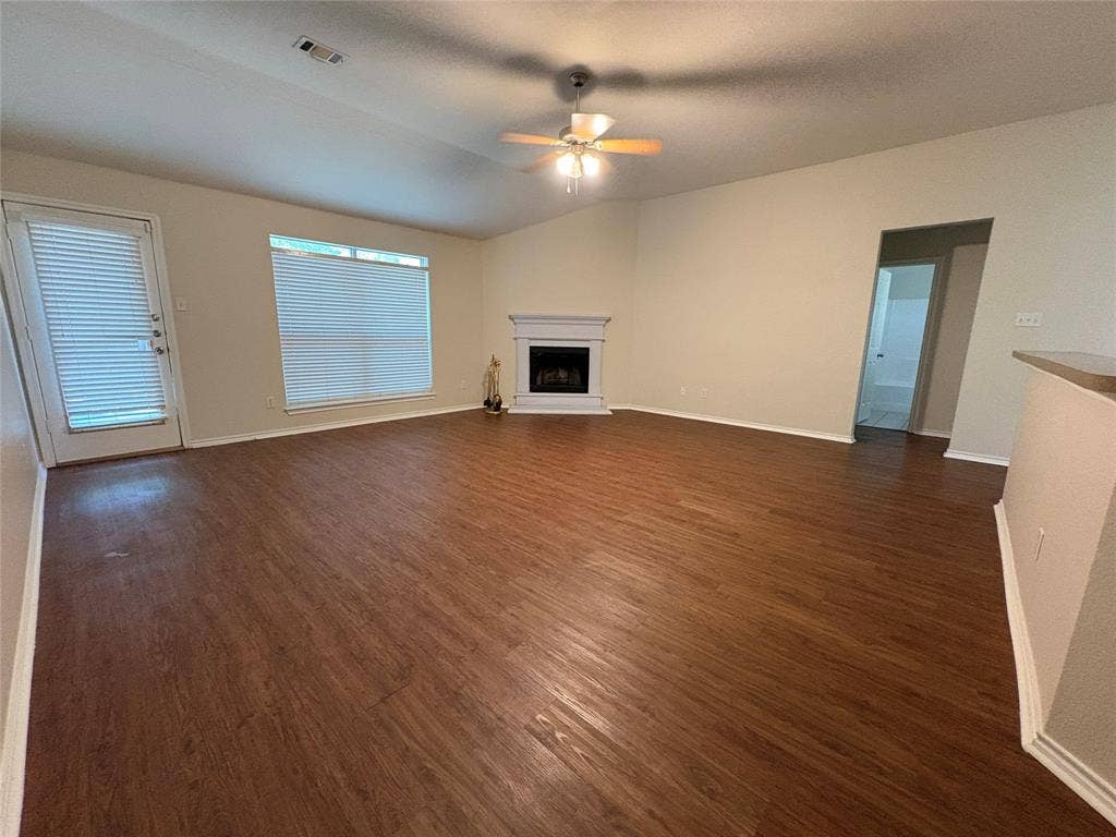 Living area featuring wood-finish flooring, a white fireplace with a decorative mantel, vaulted ceilings, a ceiling fan with integrated lighting, and a glass-paneled exterior door