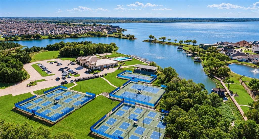 Aerial perspective of lakefront recreational facilities featuring multiple fenced tennis courts, an outdoor swimming pool, a clubhouse, and extensive paved parking