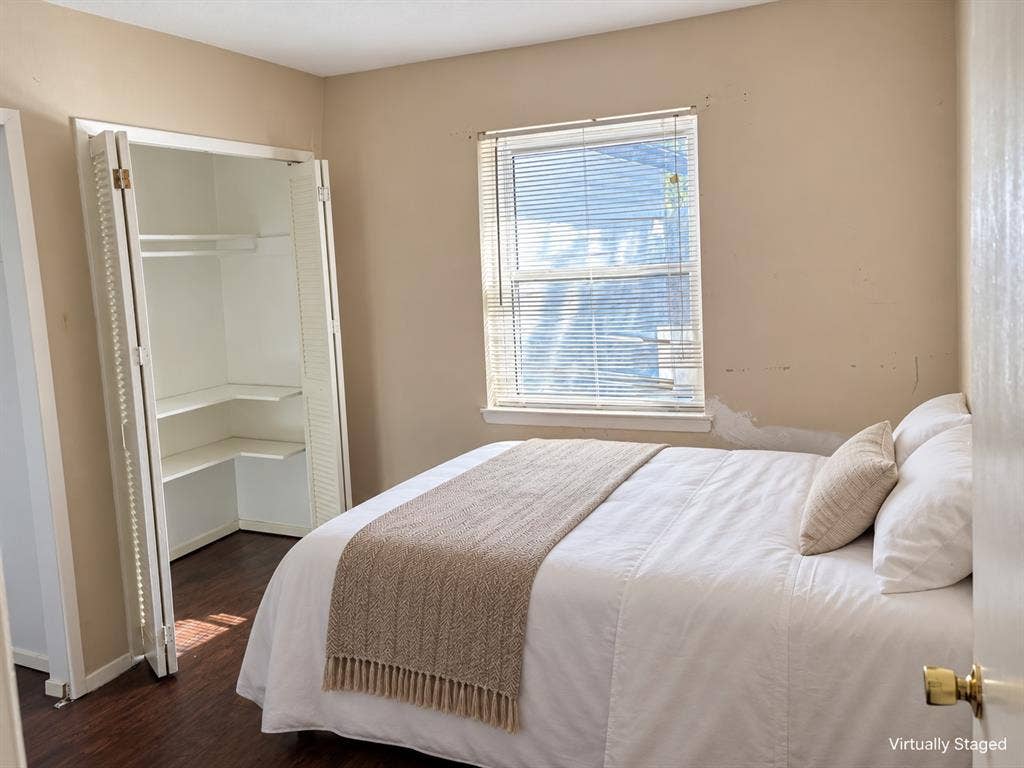 Room featuring dark wood-finish flooring, a double-hung window with built-in blinds, and a closet with bifold louvered doors and interior shelving