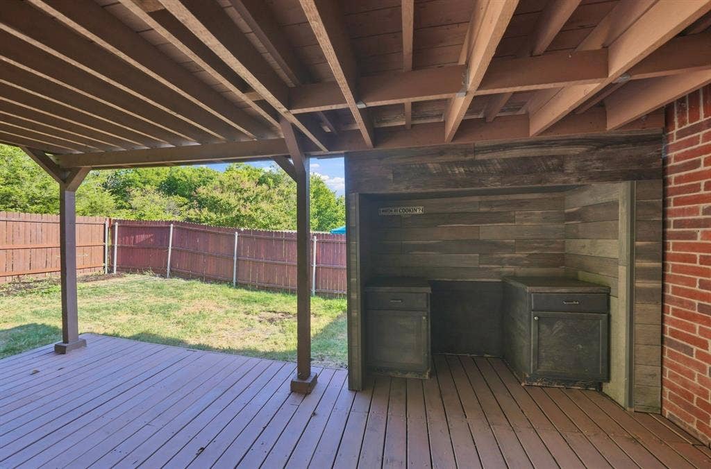 Covered wooden deck featuring an outdoor kitchen station with built-in cabinetry, wood-paneled backsplash, and brick exterior wall finish