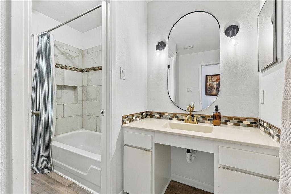 1955 Shorewood Drive, Grapevine, TX 76051 Bathroom featuring a vanity with a solid-surface countertop, an integrated sink, polished metal hardware, an oval wall mirror, and a tiled shower surround with a decorative glass mosaic inlay