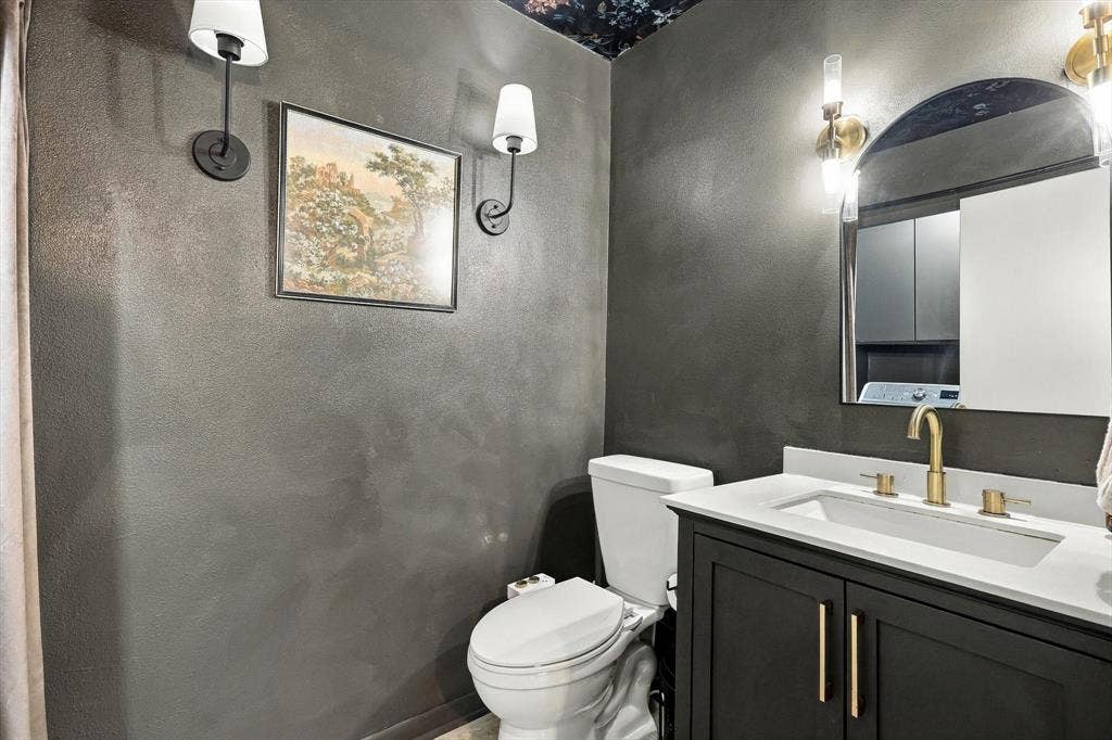 1955 Shorewood Drive, Grapevine, TX 76051 Dark vanity with a light-toned countertop, integrated basin, and brass-finish hardware, paired with dual wall-mounted light fixtures and an arched mirror