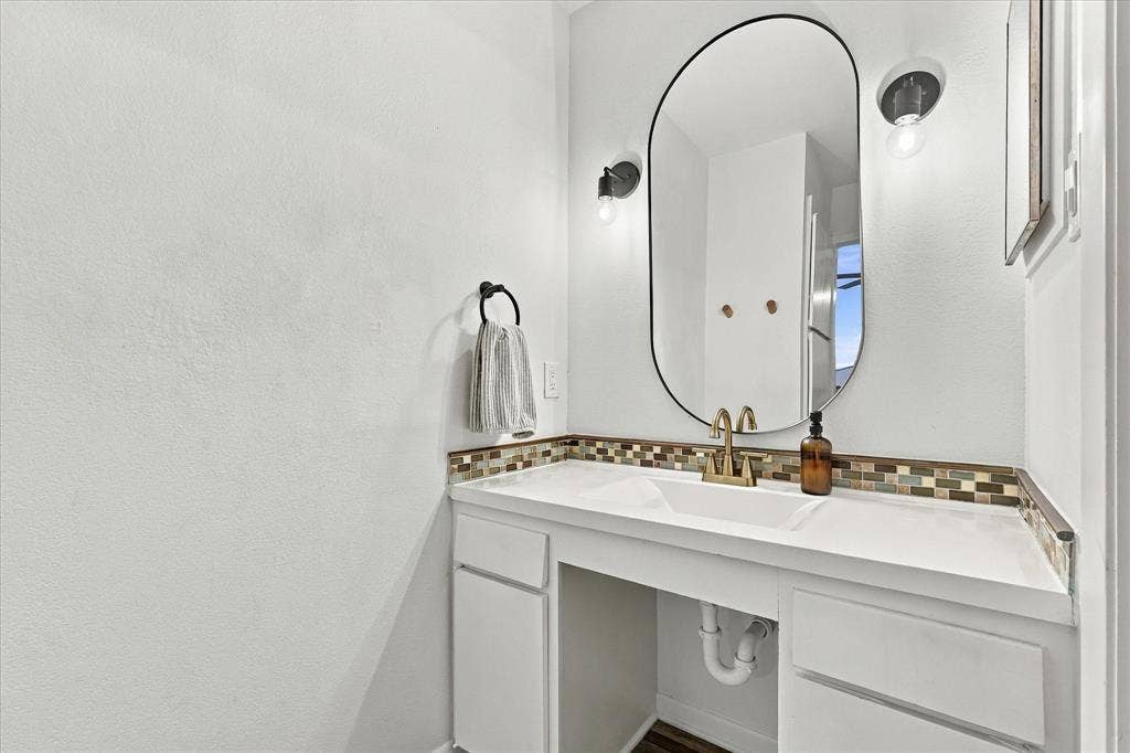 1955 Shorewood Drive, Grapevine, TX 76051 Vanity featuring an integrated sink, quartz countertop, multi-tonal mosaic tile backsplash, pill-shaped mirror, and dual wall-mounted light fixtures