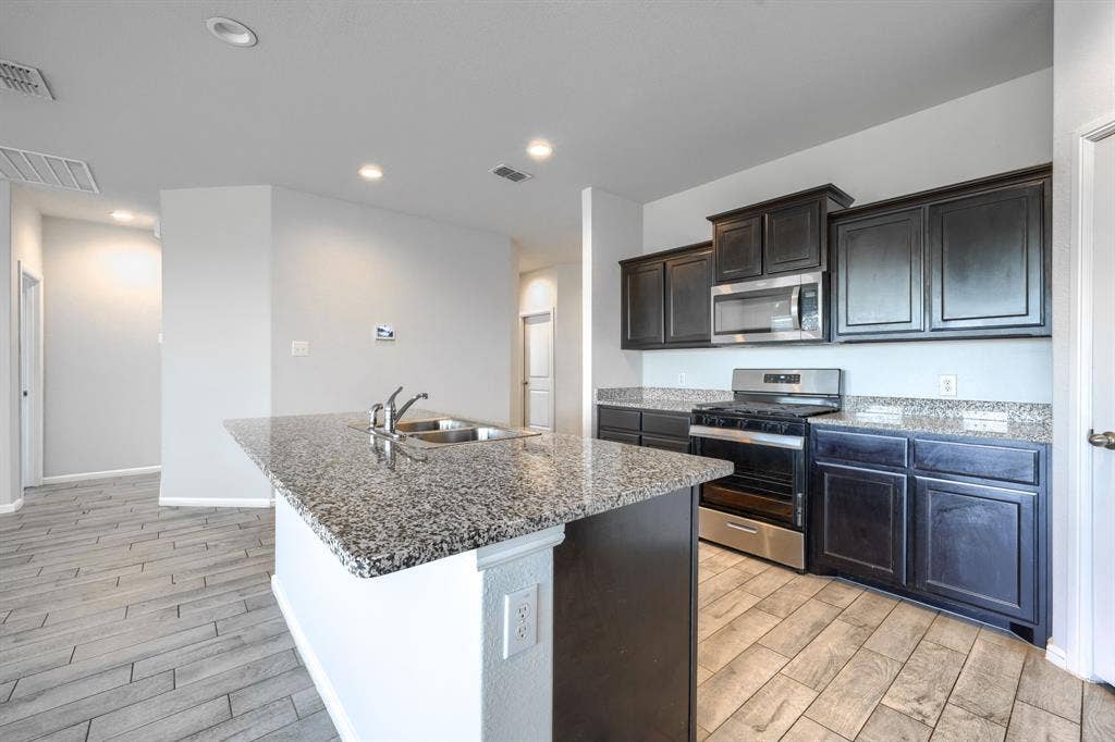 Kitchen featuring dark cabinetry, stone-finish countertops, stainless steel appliances, a center island with a dual-basin sink, and wood-finish flooring