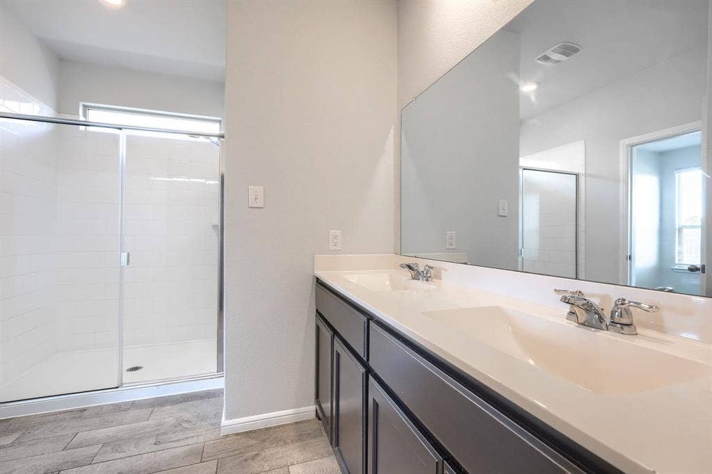 Dual-sink vanity with a solid-surface countertop, full-width mirror, glass-enclosed shower, and wood-finish tile flooring