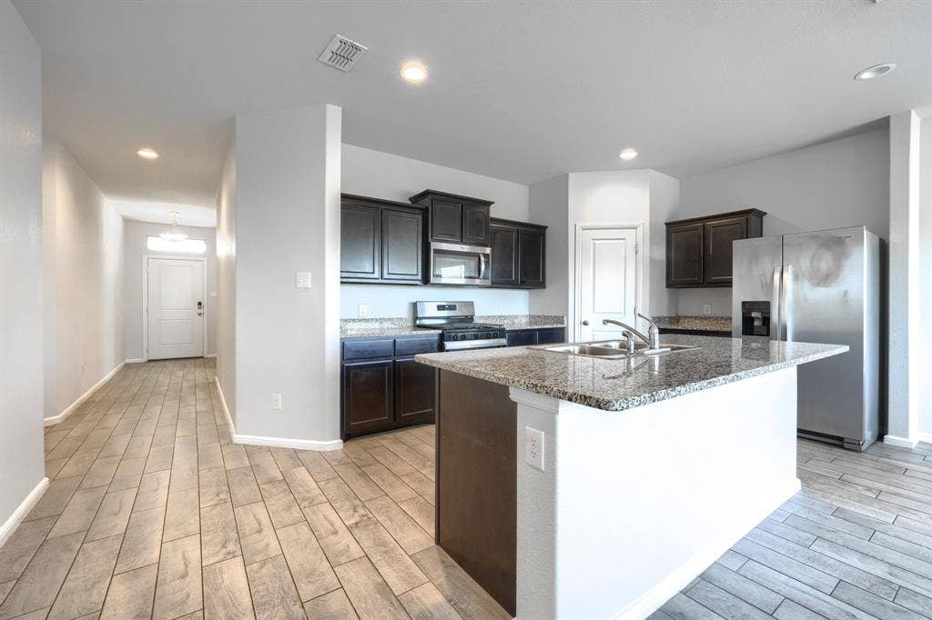 Kitchen featuring dark cabinetry, stone-finish countertops, stainless steel appliances, a center island with an integrated sink, and wood-finish flooring