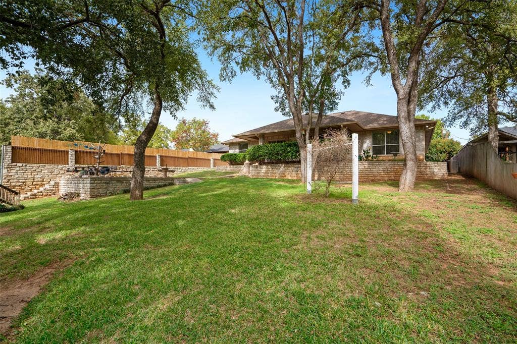 511 Casas Del Norte Court, Granbury, TX 76049 Backyard featuring a tiered stone retaining wall, mature trees, and a manicured lawn
