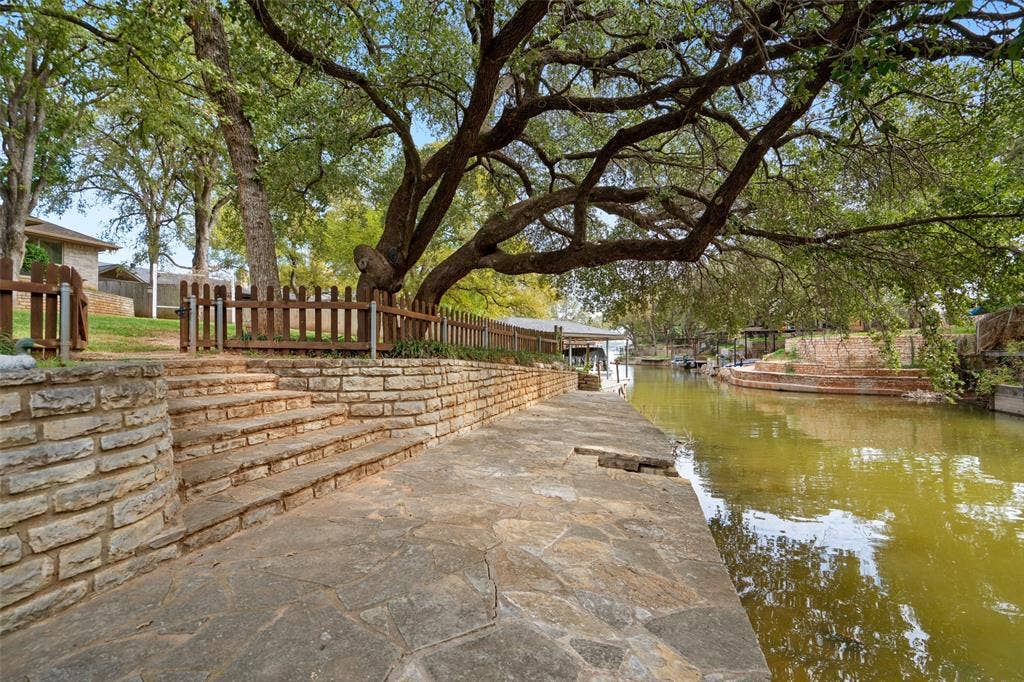 511 Casas Del Norte Court, Granbury, TX 76049 Waterfront hardscape featuring natural stone paving, terraced stone retaining walls, integrated stone stairs, and wood-slat fencing beneath mature canopy trees