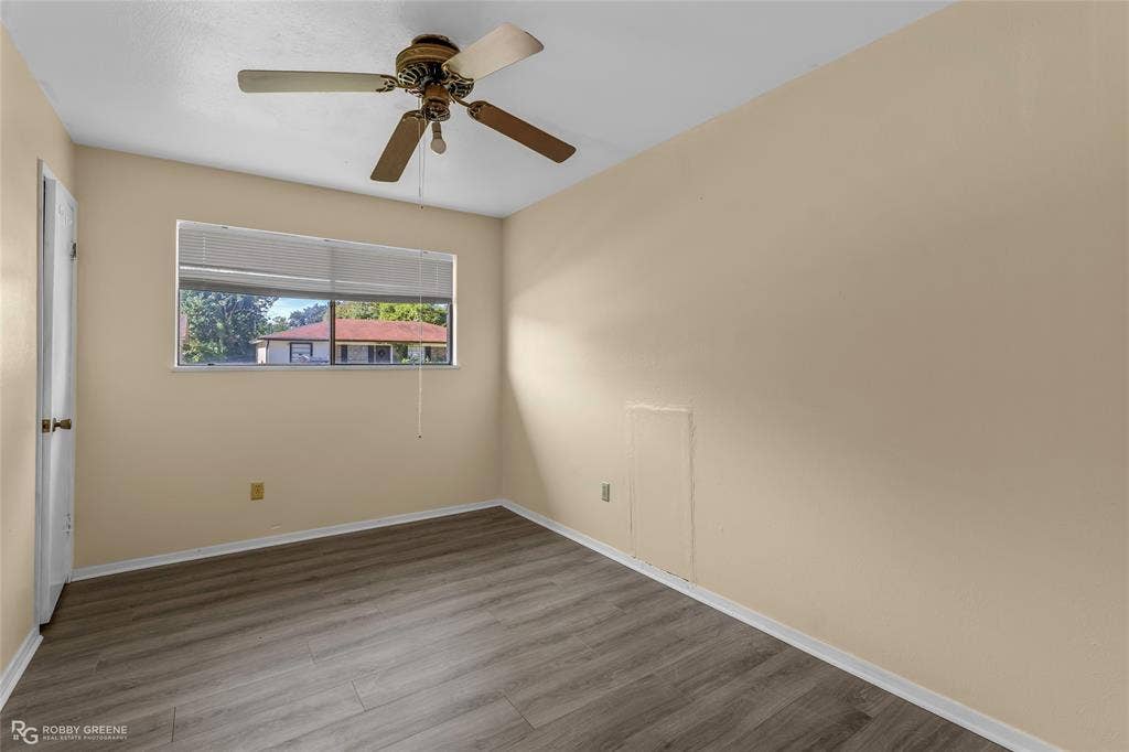Neutral-toned interior space featuring wood-finish flooring, a ceiling fan with integrated light fixture, and a horizontally oriented window with blind